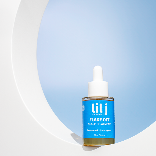A bottle of Flake Off Scalp Treatment by lil'j, positioned in the foreground with a white background and a blue sky in the background.
